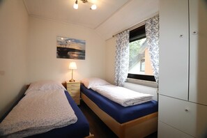Ein kleines, gut beleuchtetes Schlafzimmer mit zwei Einzelbetten, einem Nachttisch mit Lampe, einem Fenster mit gemusterten Vorhängen und einem Bild an der Wand.