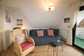 Ein gemütliches Zimmer mit einem Korbstuhl, einem grauen Sofa, einem kleinen Tisch und einem Fenster mit Vorhängen.