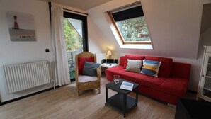 Ein Zimmer mit einem roten Sofa, einem Korbstuhl, einem kleinen Tisch und einem Fenster mit Blick auf Grünflächen.