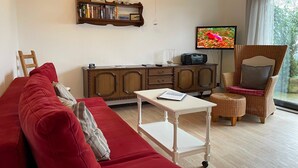 A living room with a red sofa, a white table, a wicker chair, a wooden cabinet, and a TV on the wall. - Ferienwohnungen Jade-blick, WH EG Rechts (Varel)