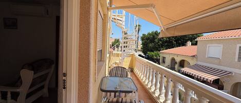 A balcony with a white balustrade, a wicker chair, a small table, and a view of neighboring buildings.