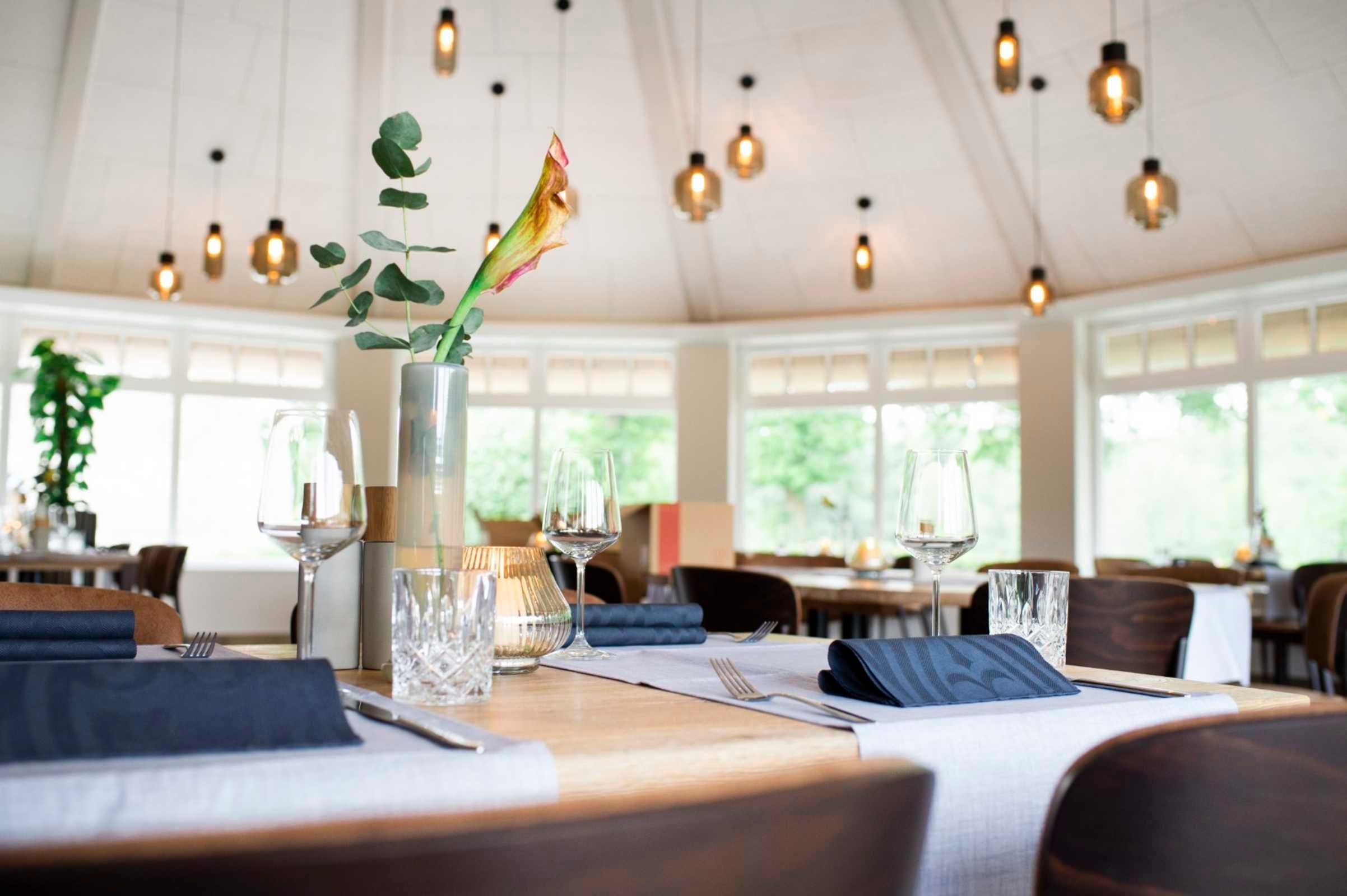 A dining area with tables set for two, a vase with a plant, and hanging pendant lights.