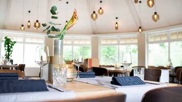 A dining area with tables set for two, a vase with a plant, and hanging pendant lights.