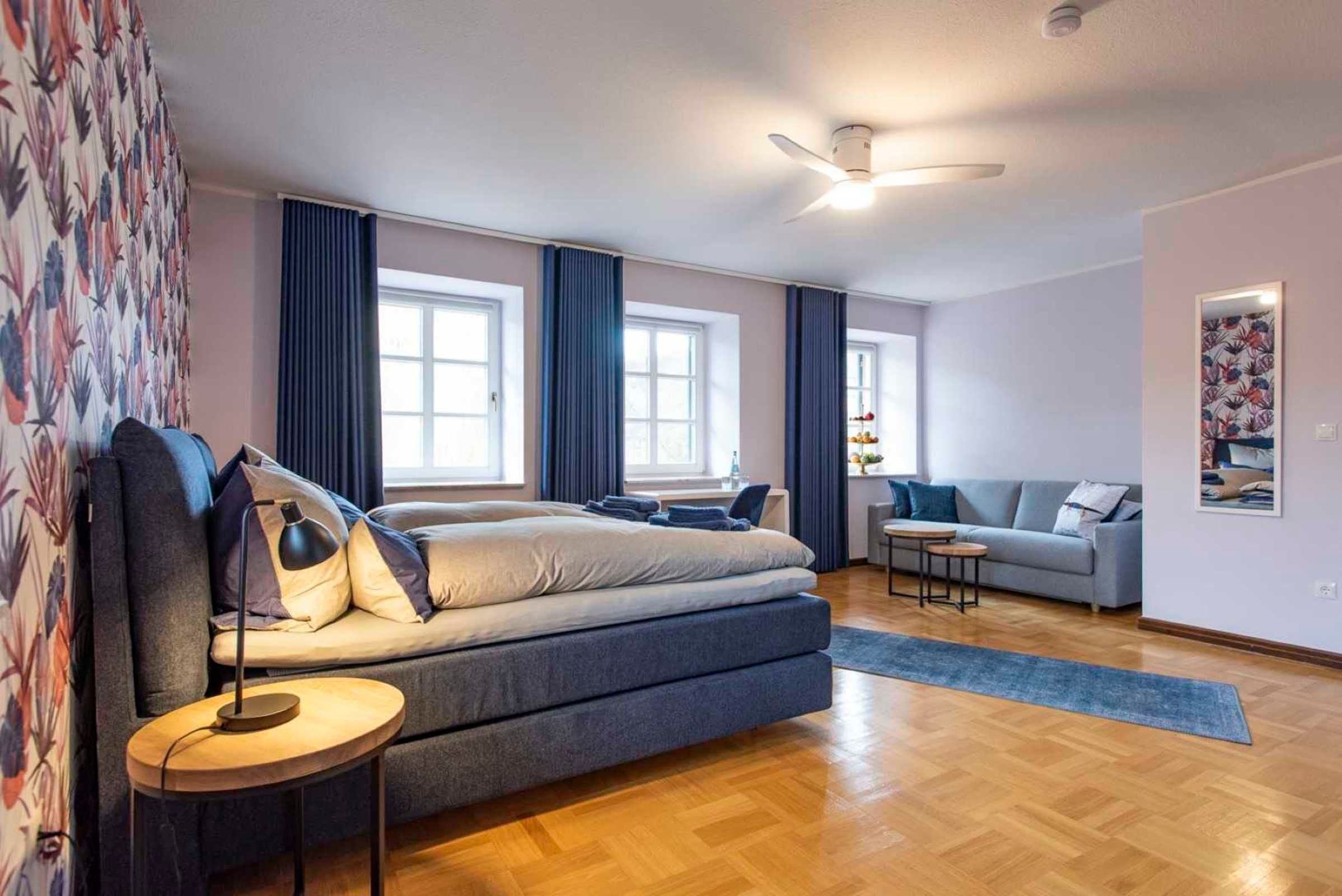 A modern bedroom with a large bed, a small table, a sofa, and a ceiling fan.