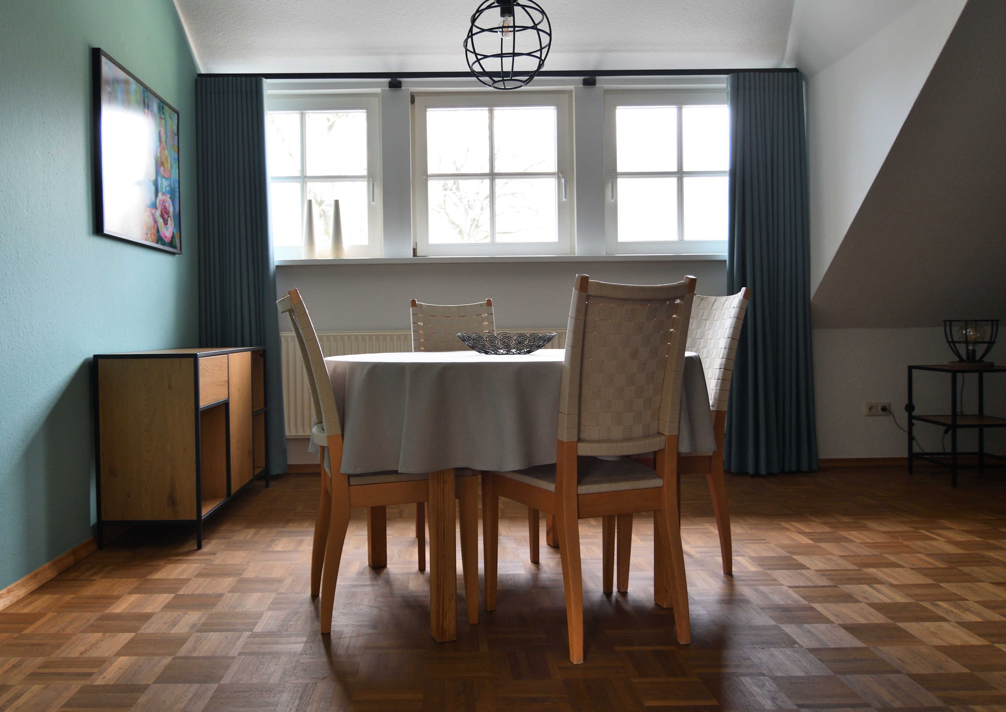 A dining area with a table set for four, wooden chairs, a radiator, and a window with curtains.