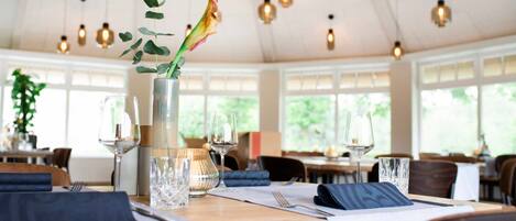 Un espace repas avec des tables préparées pour deux personnes, un vase avec une plante et des lampes suspendues.