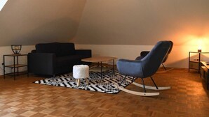 A loft-style room with a black sofa, a blue rocking chair, a small white stool, and a patterned rug on a wooden floor.