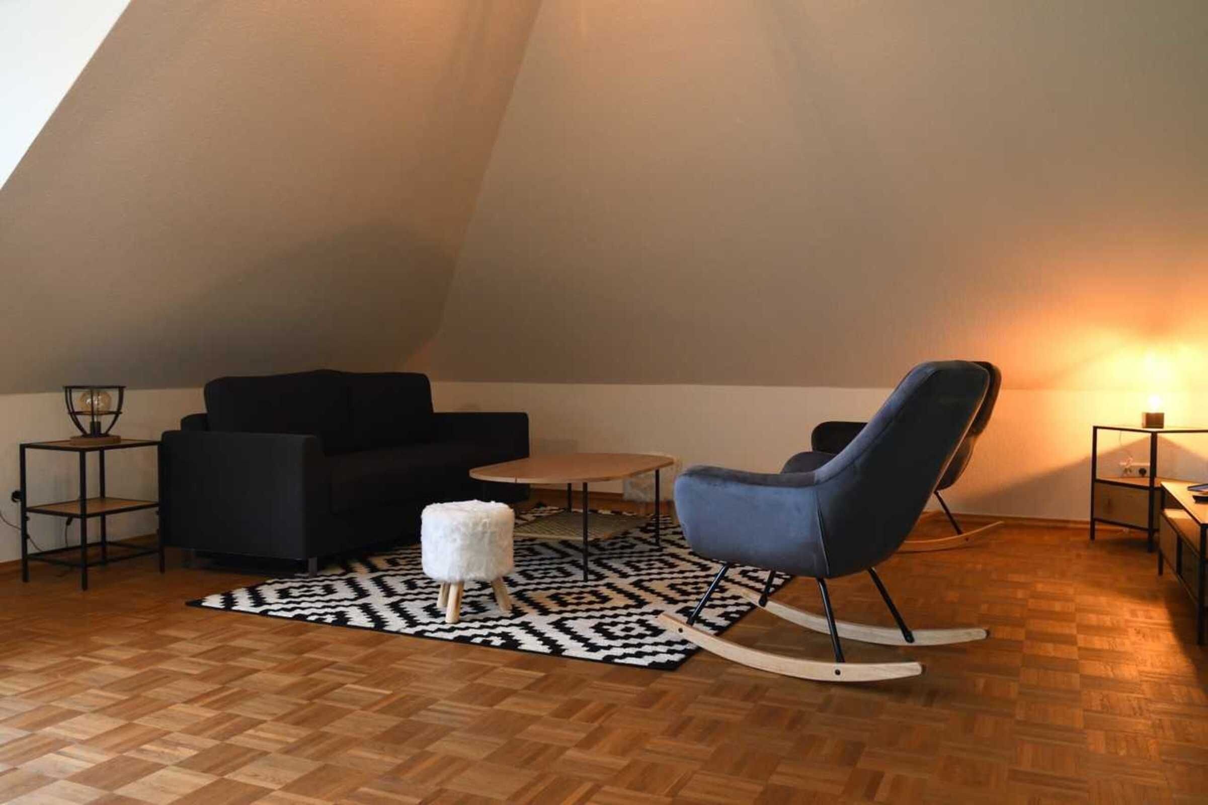 Une pièce de style loft avec un canapé noir, un fauteuil à bascule bleu, un petit tabouret blanc et un tapis à motifs sur un plancher en bois.