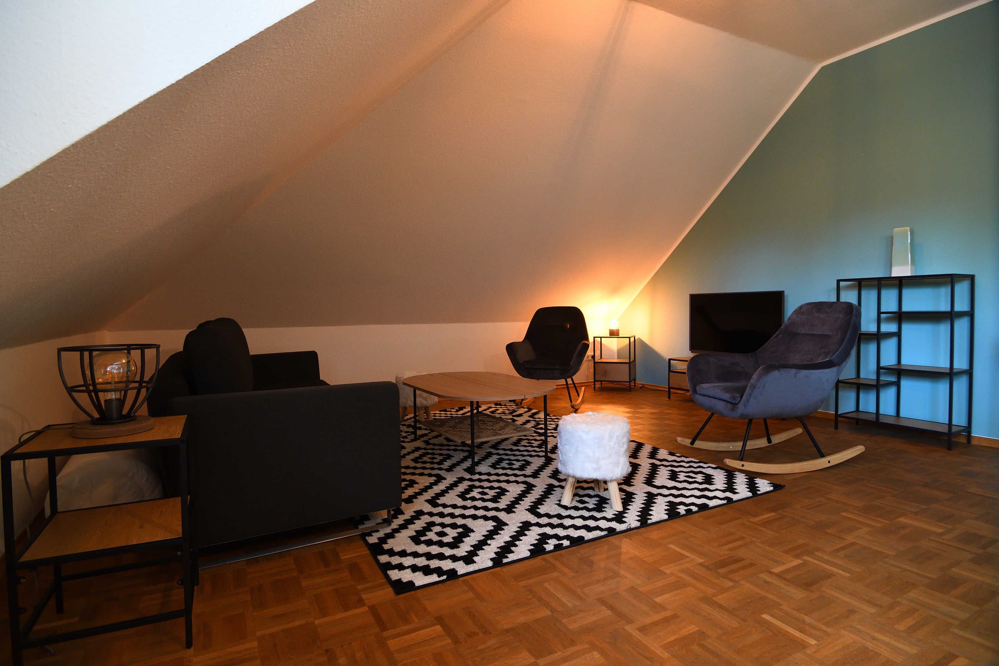 Ein Dachzimmer mit Holzboden, einer schwarzen Couch, einem weißen Hocker, einem Schaukelstuhl, einem kleinen Tisch mit Lampe und einem Bücherregal.