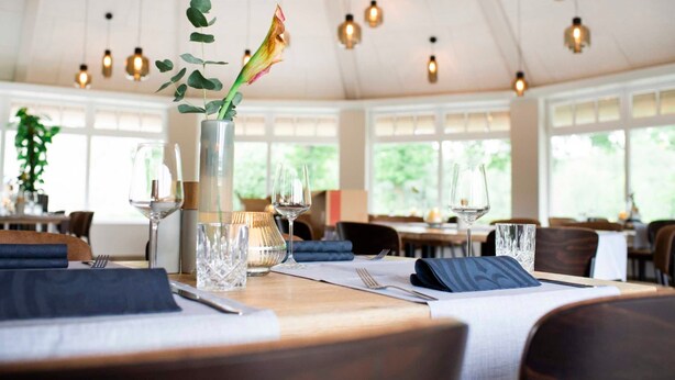 A dining area with tables set for two, a vase with a plant, and hanging pendant lights.
