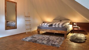 A loft bedroom with a sloped ceiling, a wooden ladder, a bed with striped bedding, a mirror, and a small table with a lamp.