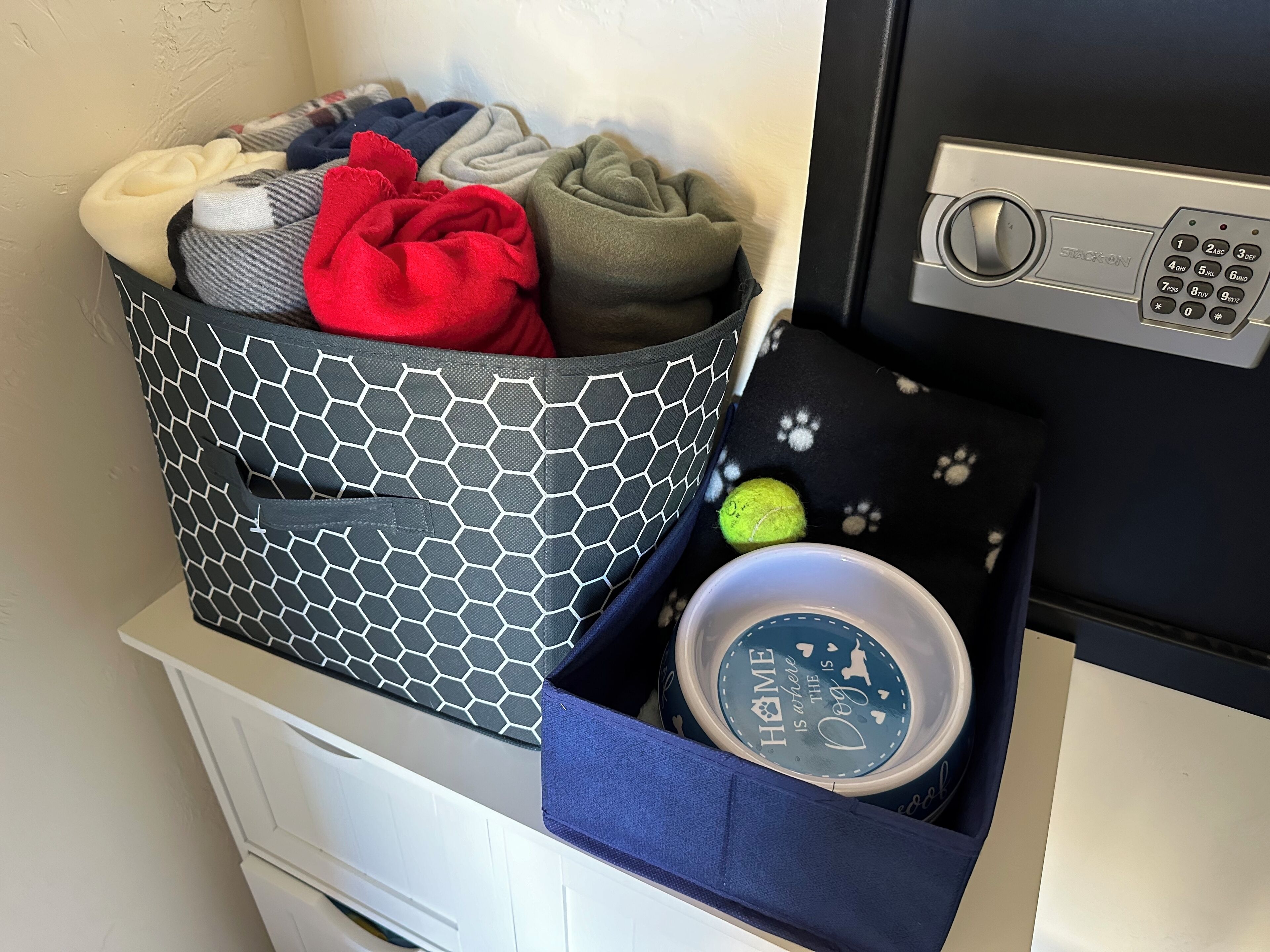 Un área de almacenamiento con una cesta estampada en blanco y negro que contiene ropa doblada, una caja azul con un diseño de huella de pata y un pequeño recipiente con la etiqueta 'HOME IS WHERE THE DOG IS'.