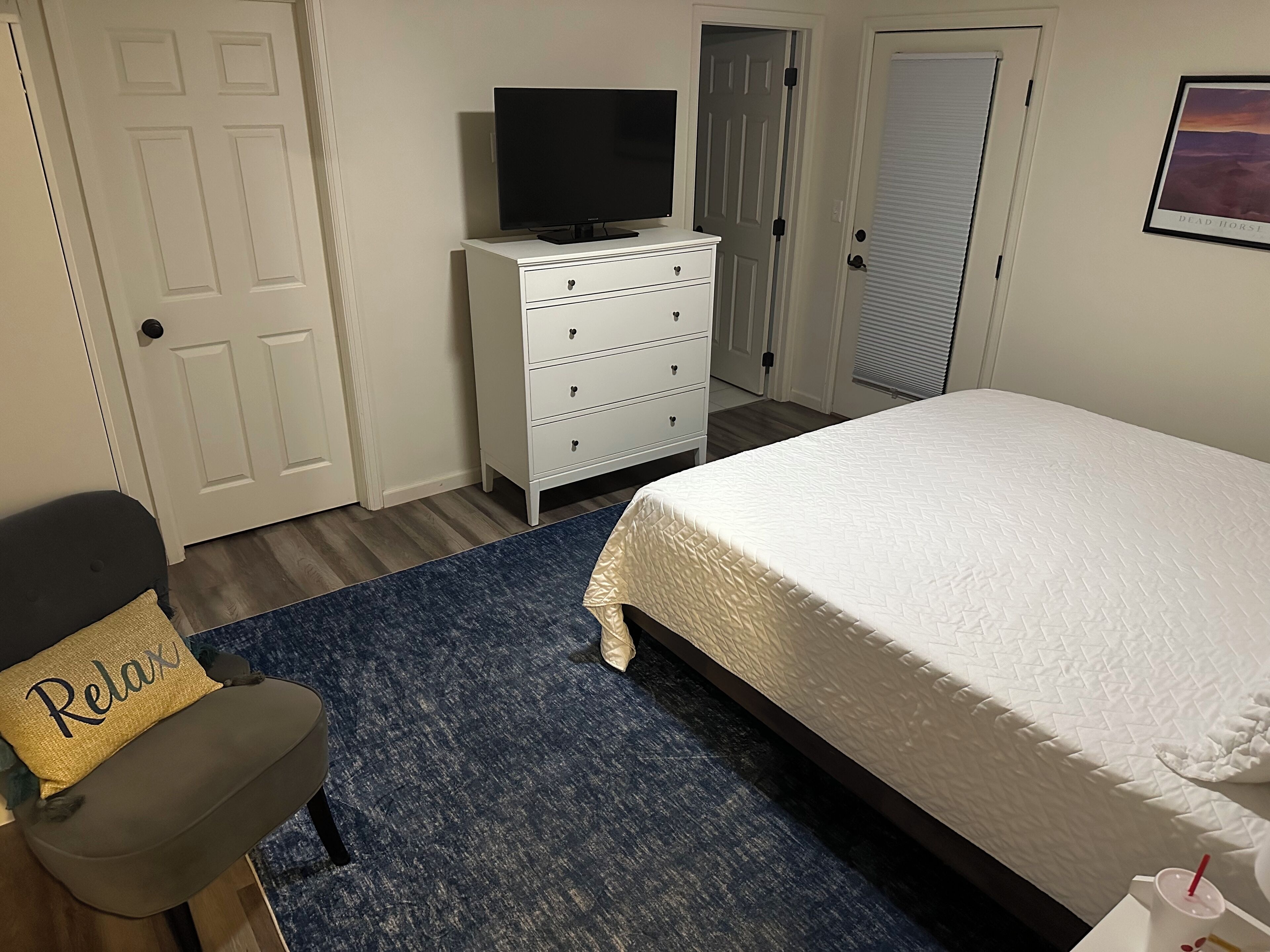 Un dormitorio con cama, televisor en una cómoda, una silla con cojín que dice 'Relax' y una alfombra azul.