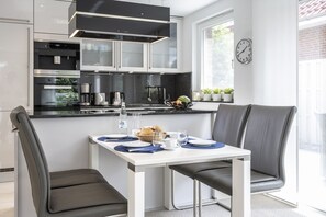 A modern kitchen with white cabinets, a black countertop, and stainless steel appliances. There is a dining table with chairs, a clock on the wall, and a window with plants.