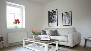 A living room with a white sofa, a wooden coffee table, a vase with flowers, and two framed botanical prints on the wall.