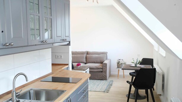 Eine moderne Küche mit integriertem Spülbecken und Schränken, ein Essbereich mit Tisch und Stühlen sowie eine gemütliche Sitzecke mit Sofa und Kissen.