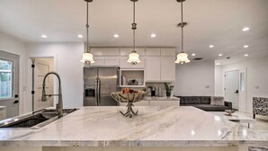 Een moderne keuken met een marmeren eiland, roestvrijstalen apparatuur, hanglampen, en een grijze bank in de woonkamer.