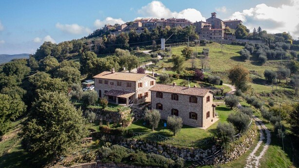 Un bâtiment en pierre, surmonté d’un toit de tuiles, est entouré de verdure. Au loin, on aperçoit une colline sur laquelle se dresse un grand bâtiment.