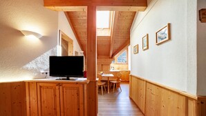 Un interior de madera con un televisor sobre un mueble, un comedor y cuadros en la pared.