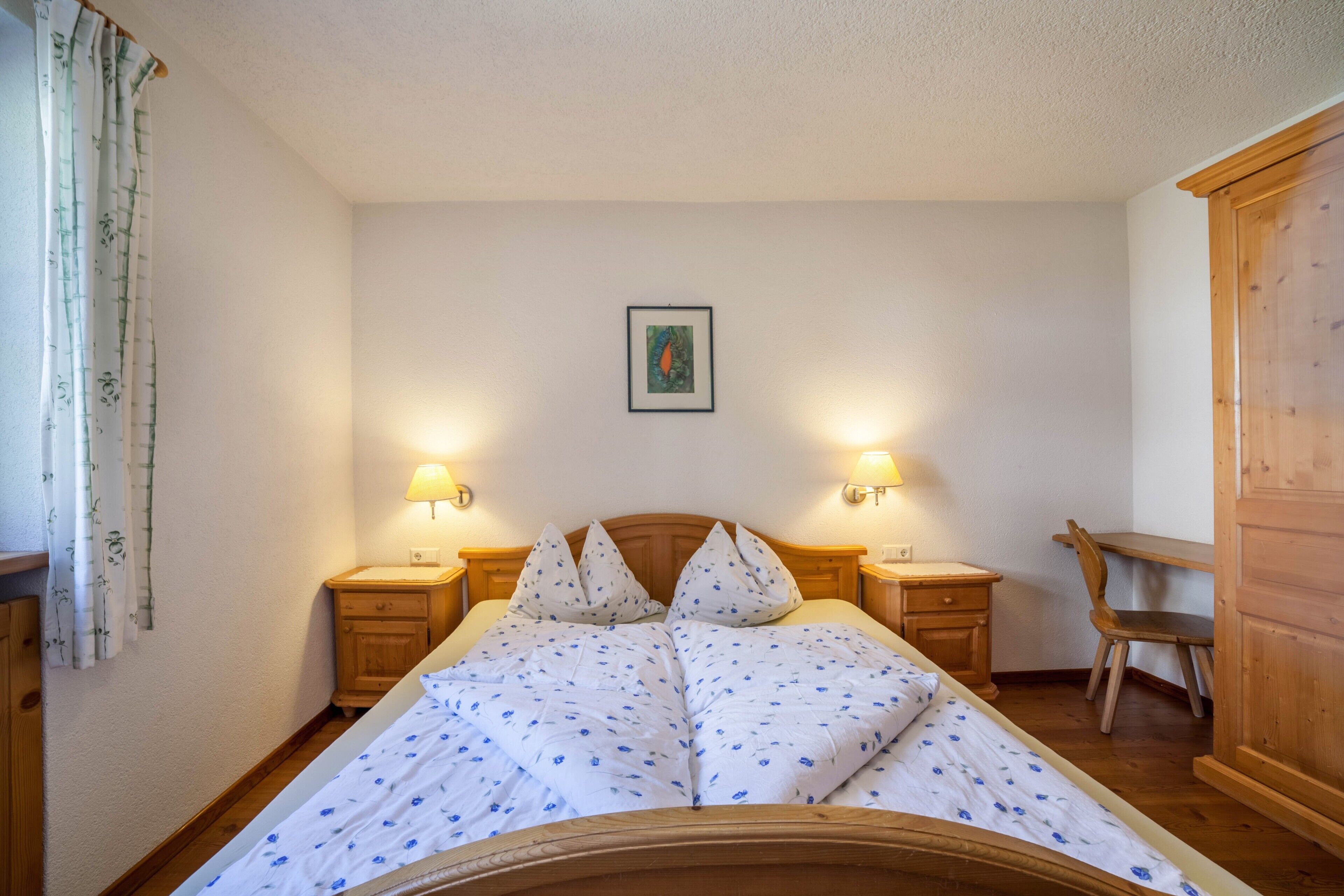 Une chambre à coucher avec un lit en bois, deux tables de chevet avec des lampes, une chaise en bois, une armoire en bois et un tableau encadré accroché au mur.