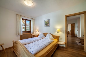 A room with a double bed, a window with curtains, a bedside table, a wall-mounted lamp, and a door leading to another room.