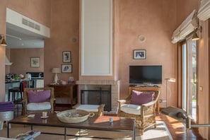 Sala de estar con muebles de mimbre, chimenea, televisor y una zona de cocina al fondo.