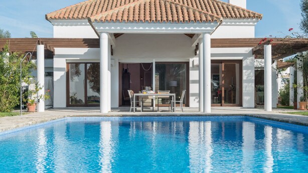 Ein Schwimmbad mit überdachter Terrasse, einem Tisch mit Stühlen und einem Gebäude mit einem Ziegeldach.