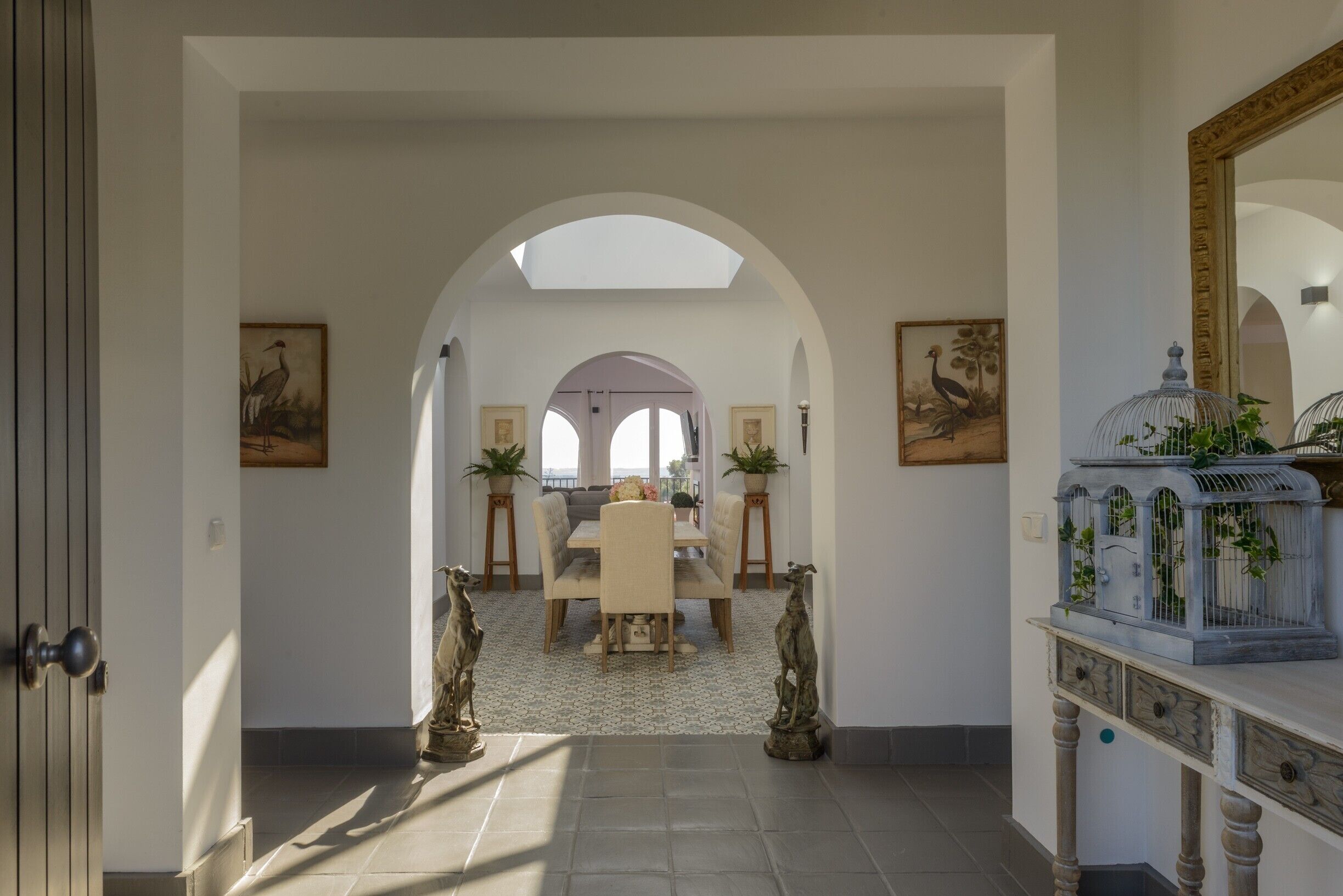 Un pasillo en arco que conduce a un comedor con una mesa, sillas, dos estatuas y una jaula de pájaros sobre una consola.