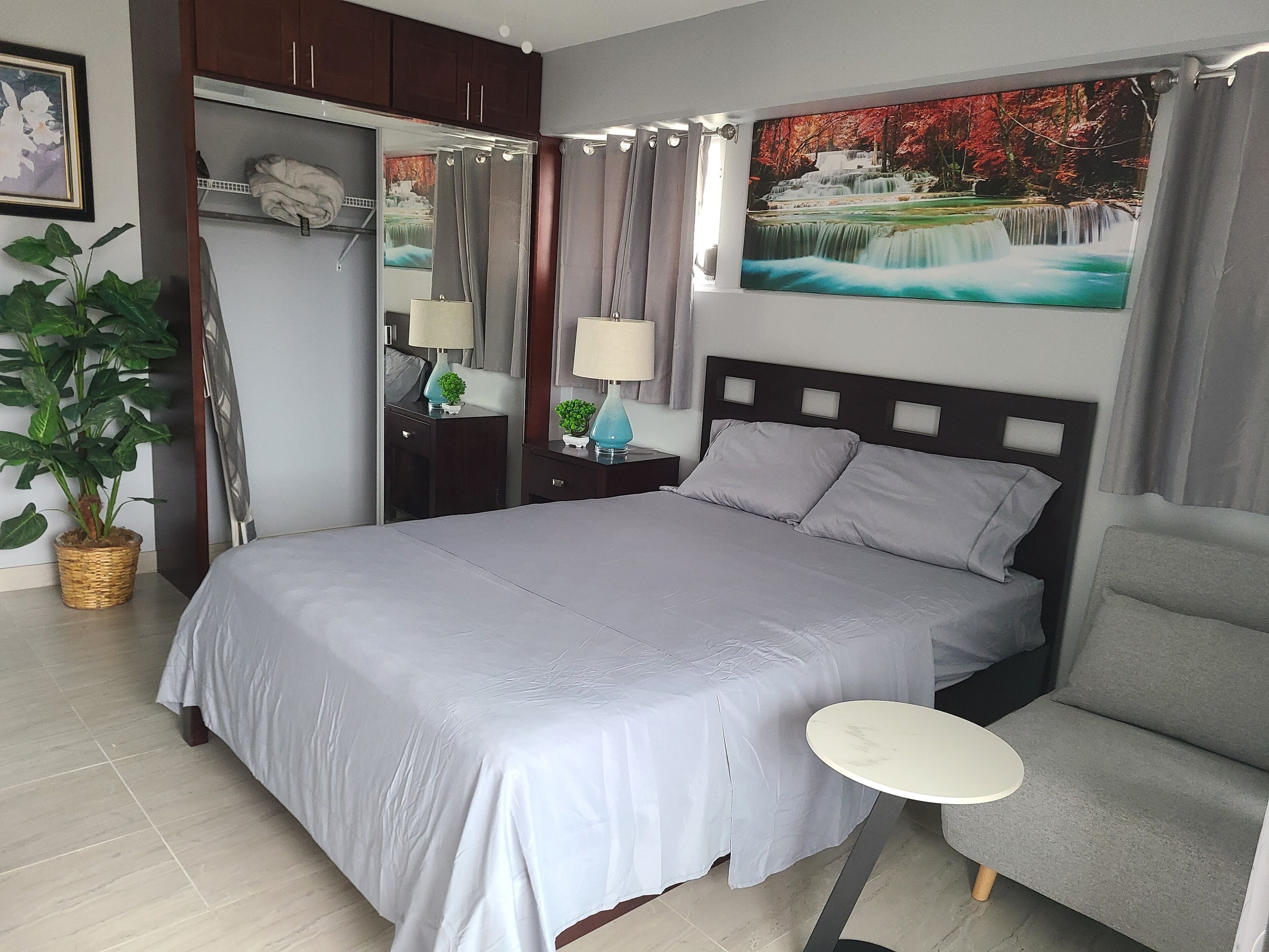 A neatly arranged bedroom with a bed, bedside tables, a chair, a small round table, a potted plant, and a painting on the wall.
