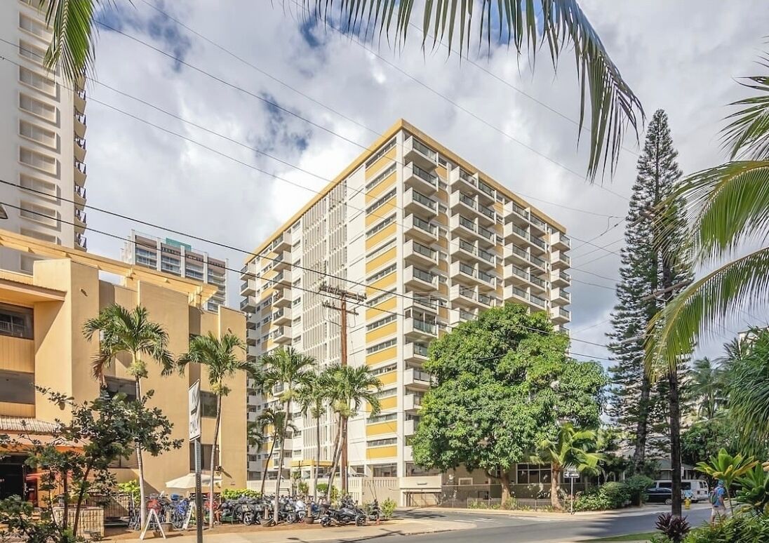 A multi-story building with balconies, surrounded by palm trees and other greenery.