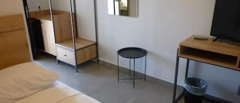 A room with a bed, a desk, a chair, a small stool, a trash bin, and a mirror.