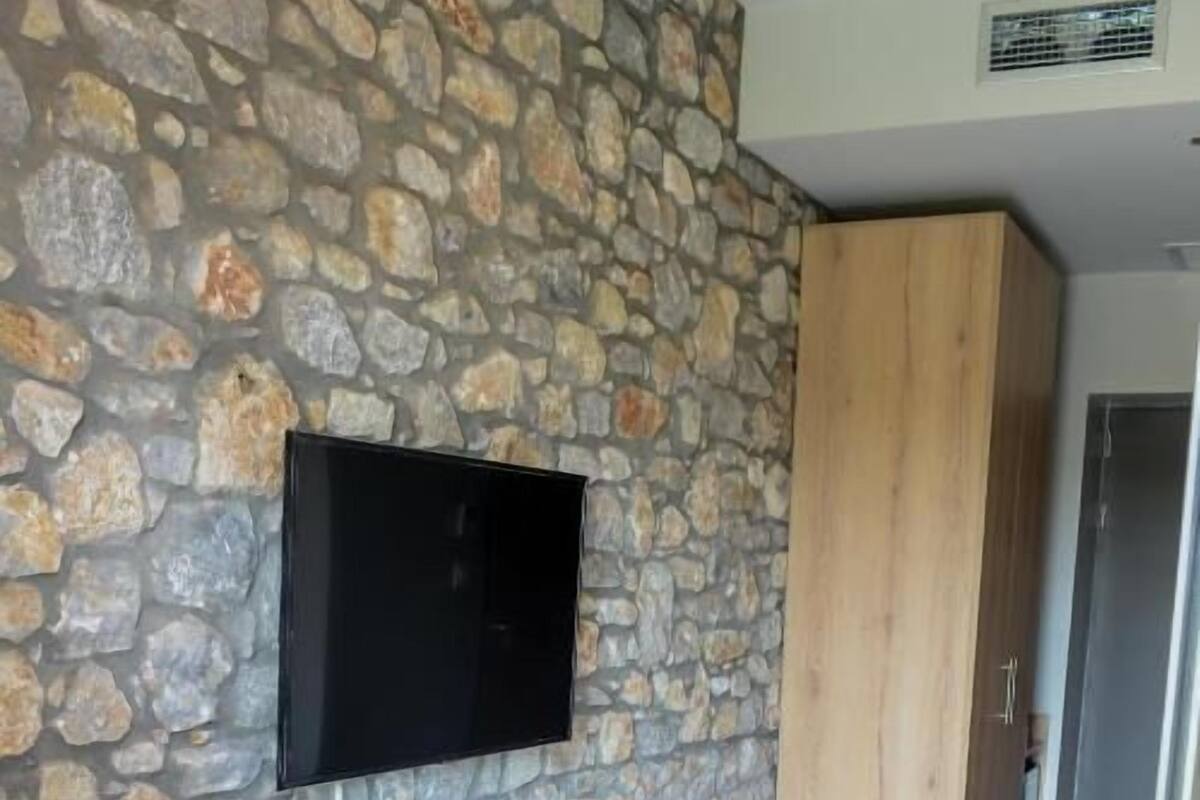 A room with a stone wall, a flat-screen TV mounted above a wooden desk, a chair, and a wardrobe.