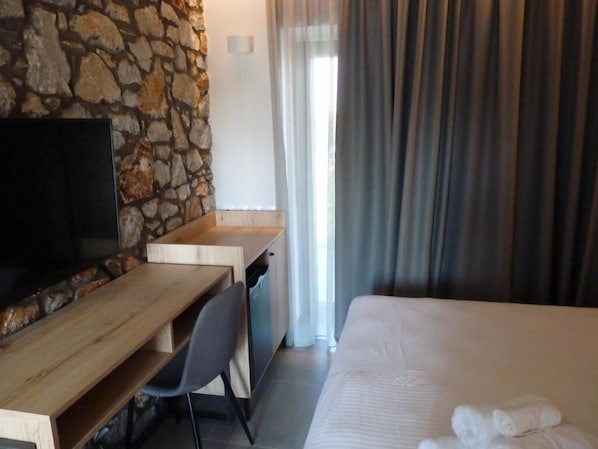 A hotel room with a stone wall, a wooden desk with a chair, a bed with white bedding, and a TV on the wall. - Comfort Suites and Rooms (Larissa)