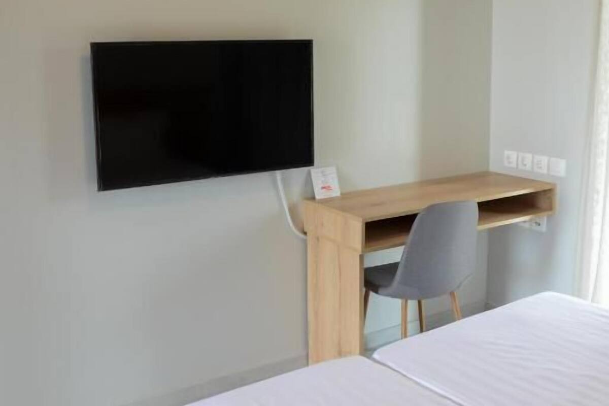 A hotel room with a bed, a desk with a chair, a flat-screen TV mounted on the wall, and a window.