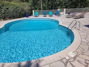A clear blue pool with a stone border, surrounded by lounge chairs and a metal railing.