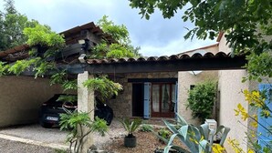 Une maison en pierre avec un toit en tuiles, une voiture garée devant, et un jardin agrémenté de plantes en pot et de fleurs jaunes.