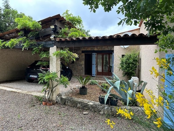 A stone house with a tiled roof, a car parked outside, and a garden with potted plants and yellow flowers.