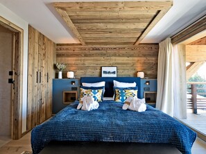 A bedroom with a wooden ceiling, a bed with blue bedding, two bedside tables with lamps, and a window with curtains.