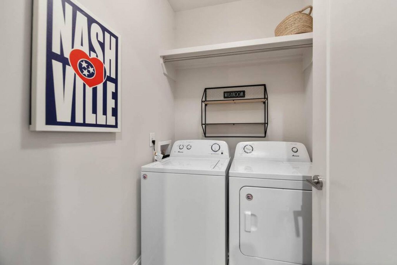 A laundry room with two front-loading washing machines, a wall-mounted sign indicating a washroom, and a framed sign with 'NASHVILLE' and a heart-shaped logo.