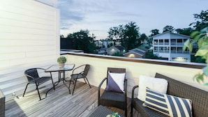 Un balcón exterior con terraza de madera, mobiliario de mimbre y vistas de edificios circundantes.