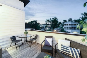 An outdoor balcony with a wooden deck, wicker furniture, and a view of surrounding buildings.