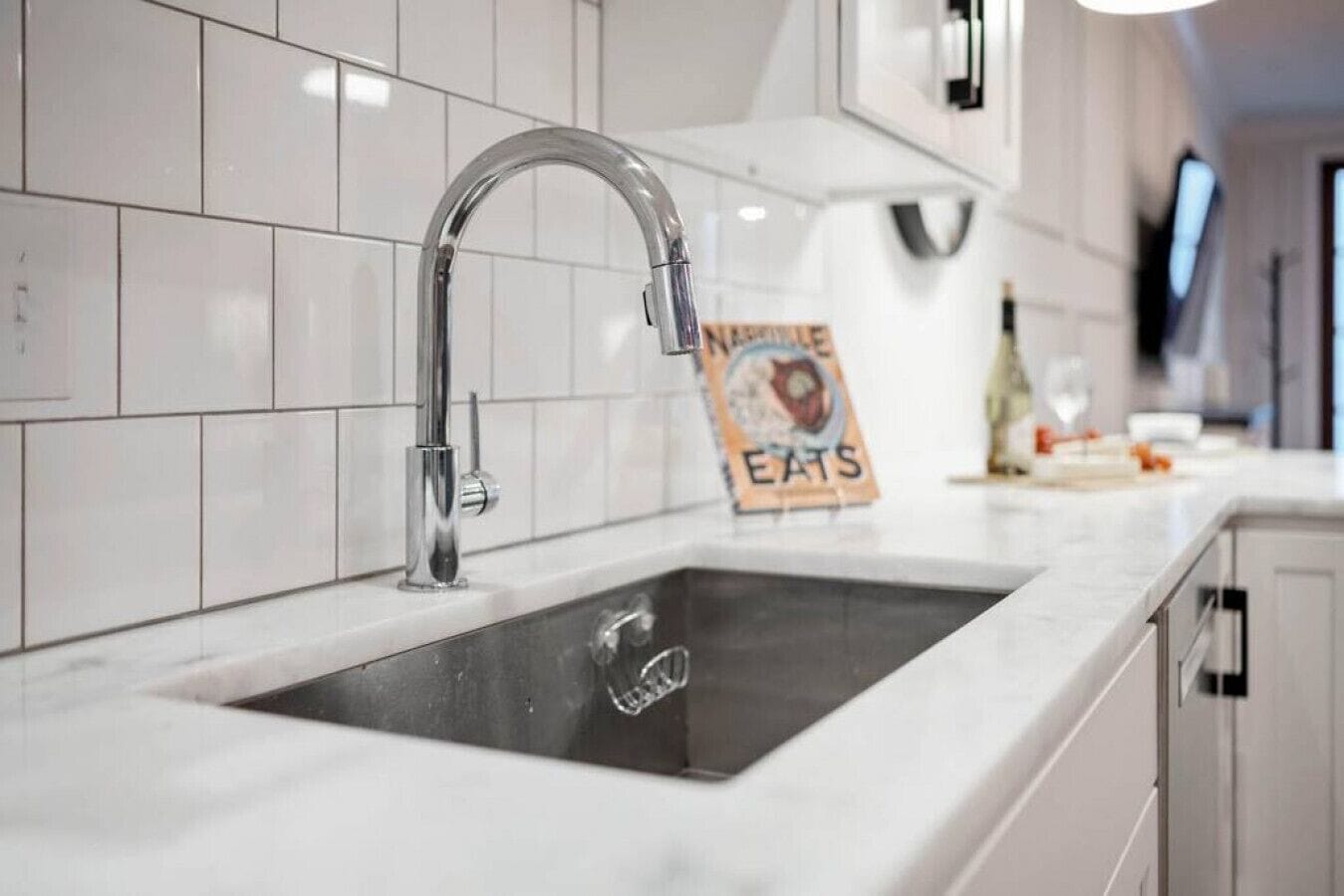 A kitchen with a stainless steel faucet, white tiled backsplash, and a sign on the counter.