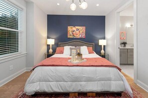 Un dormitorio con cama, lámparas de noche, un cuadro en la pared y vistas al baño.