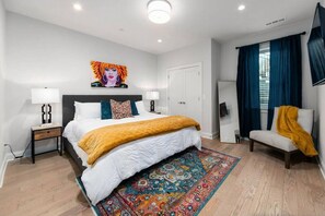 A modern bedroom with a large bed, a nightstand, a bedside table, a chair, a mirror, and a colorful rug.