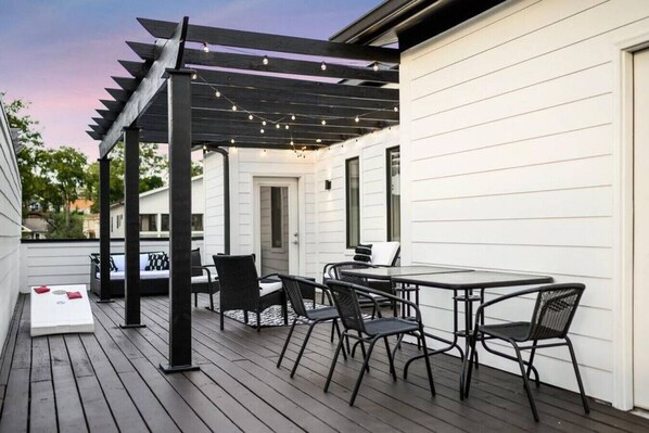 A covered outdoor patio with a table and chairs, string lights, and a view of a white building.