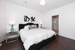 A modern bedroom with a large bed, two bedside tables with lamps, a dark wood floor, and a glass shower door.