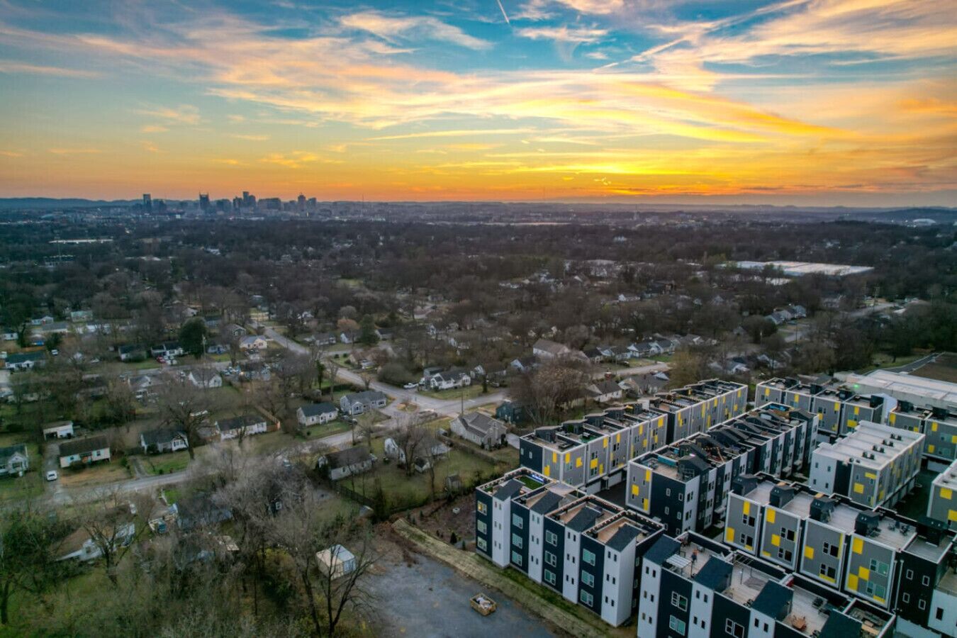 A residential area with modern apartment buildings, a mix of single and multi-story houses, and a distant city skyline during sunset.