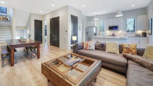 Una sala de estar moderna con una mesa de centro de madera, un sofá con cojines estampados y una zona de cocina con refrigerador y microondas.