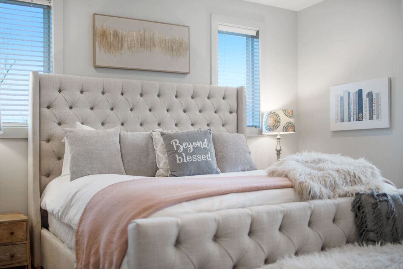 Un dormitorio con cabecera capitonada, cama con manta rosa y una manta mullida. Hay dos ventanas con persianas, una mesita de noche y una lámpara.