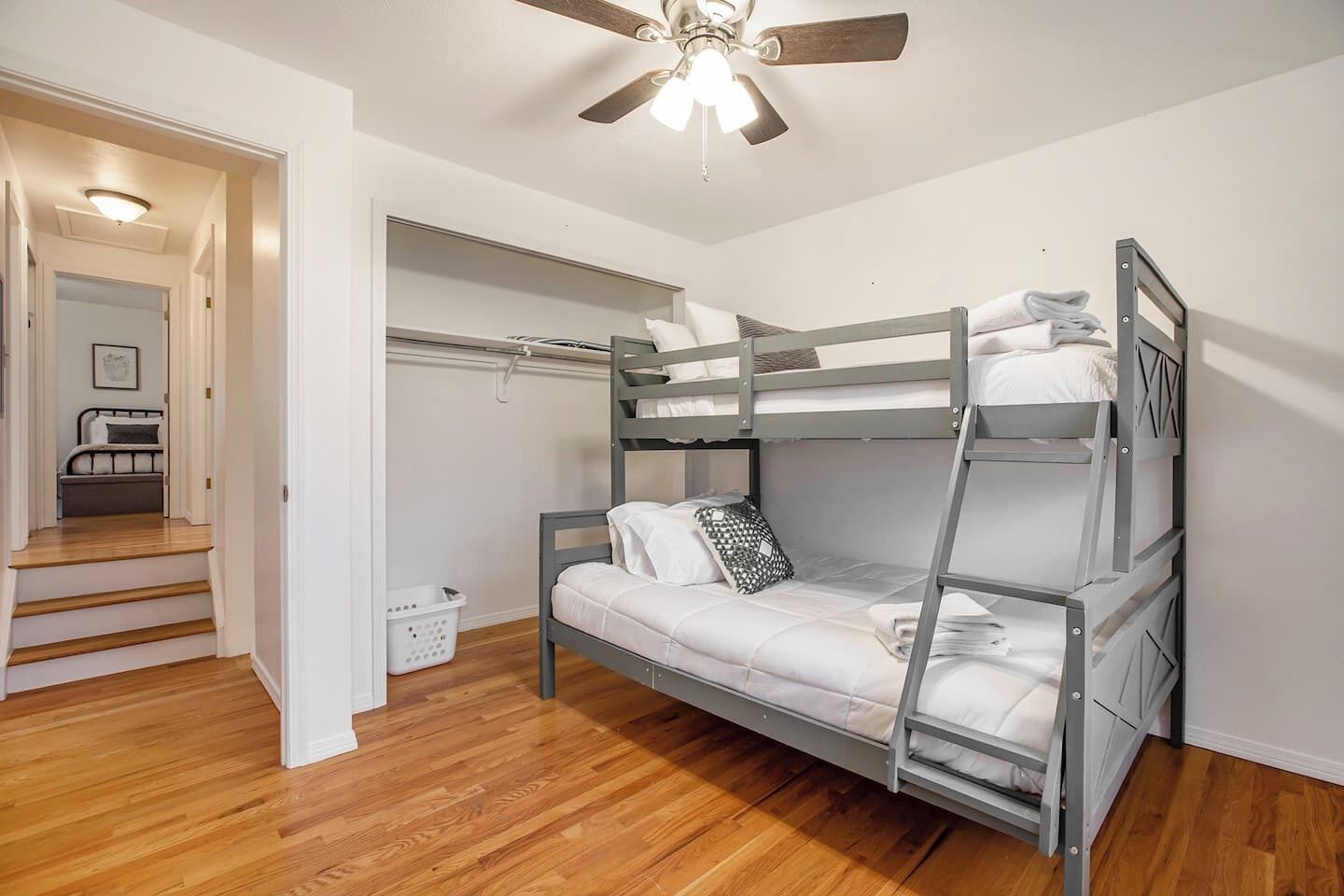 A bunk bed room with a ceiling fan, wooden flooring, and a hallway leading to another room.